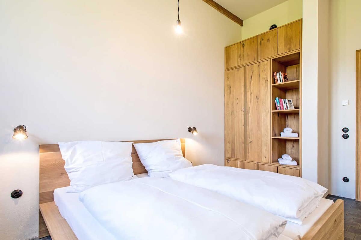 Un lit double avec du linge de lit blanc, une tête de lit en bois, deux lampes de chevet et une armoire en bois avec des étagères.