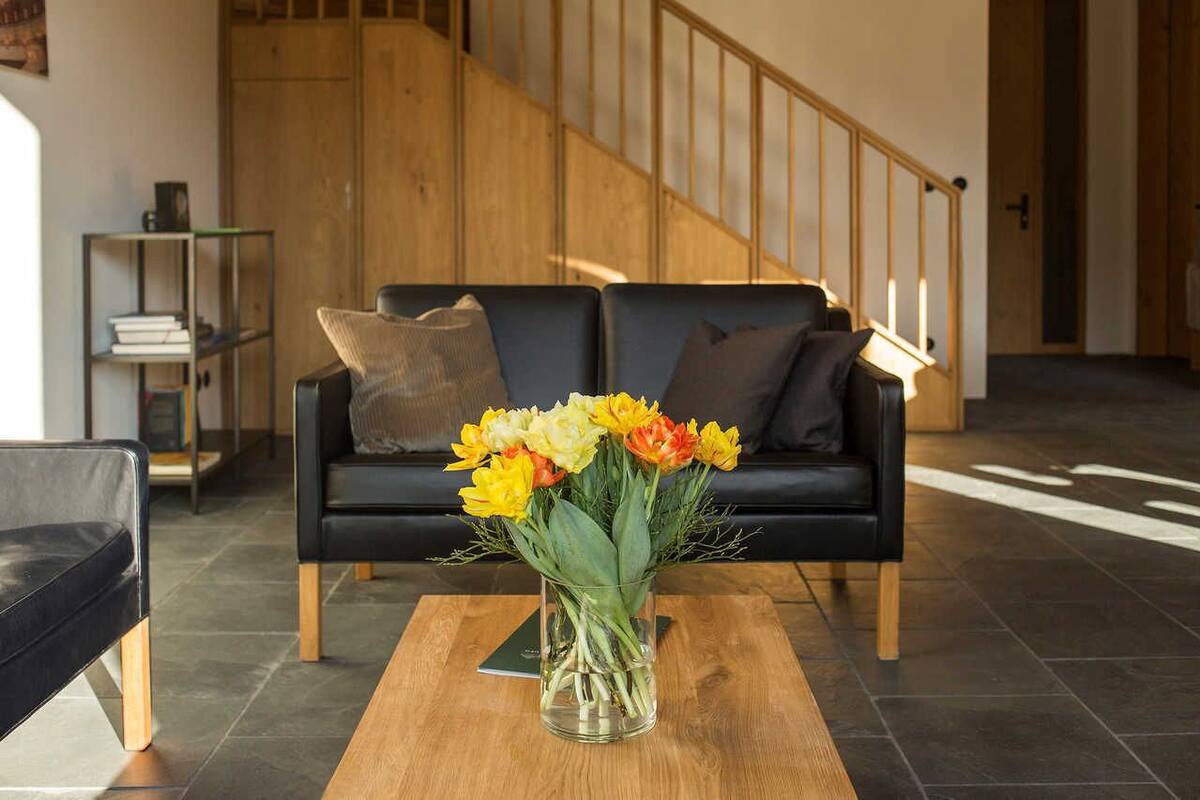 Un salon moderne avec un canapé en cuir noir, un vase en verre rempli de fleurs et un escalier en bois en arrière-plan.