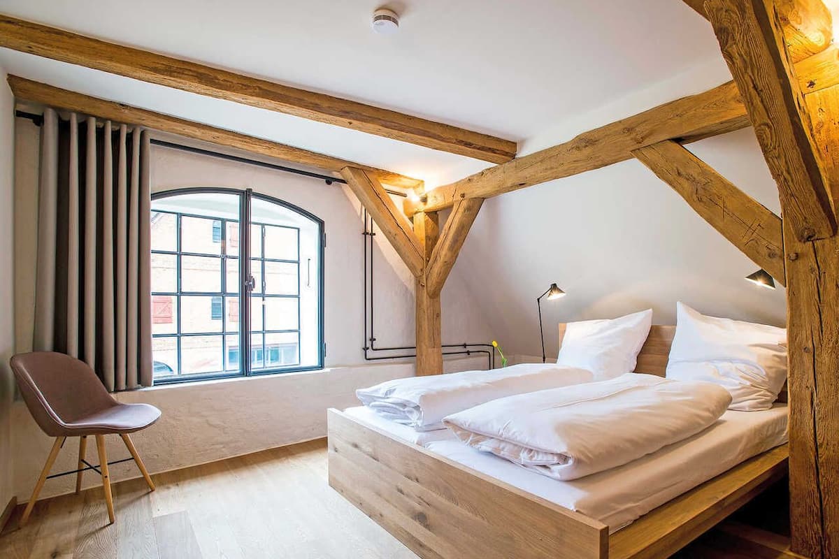 Une chambre avec un plafond en bois, une grande fenêtre avec des rideaux, un lit avec du linge de lit blanc et une chaise.