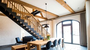 A modern interior with a wooden staircase, a dining area with a long table and chairs, and a large window providing natural light.