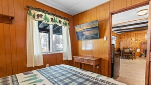 Ein Zimmer mit Holzwänden, einem Bett mit gesteppter Decke, einem an der Wand montierten Fernseher und einer Tür zu einem Essbereich.