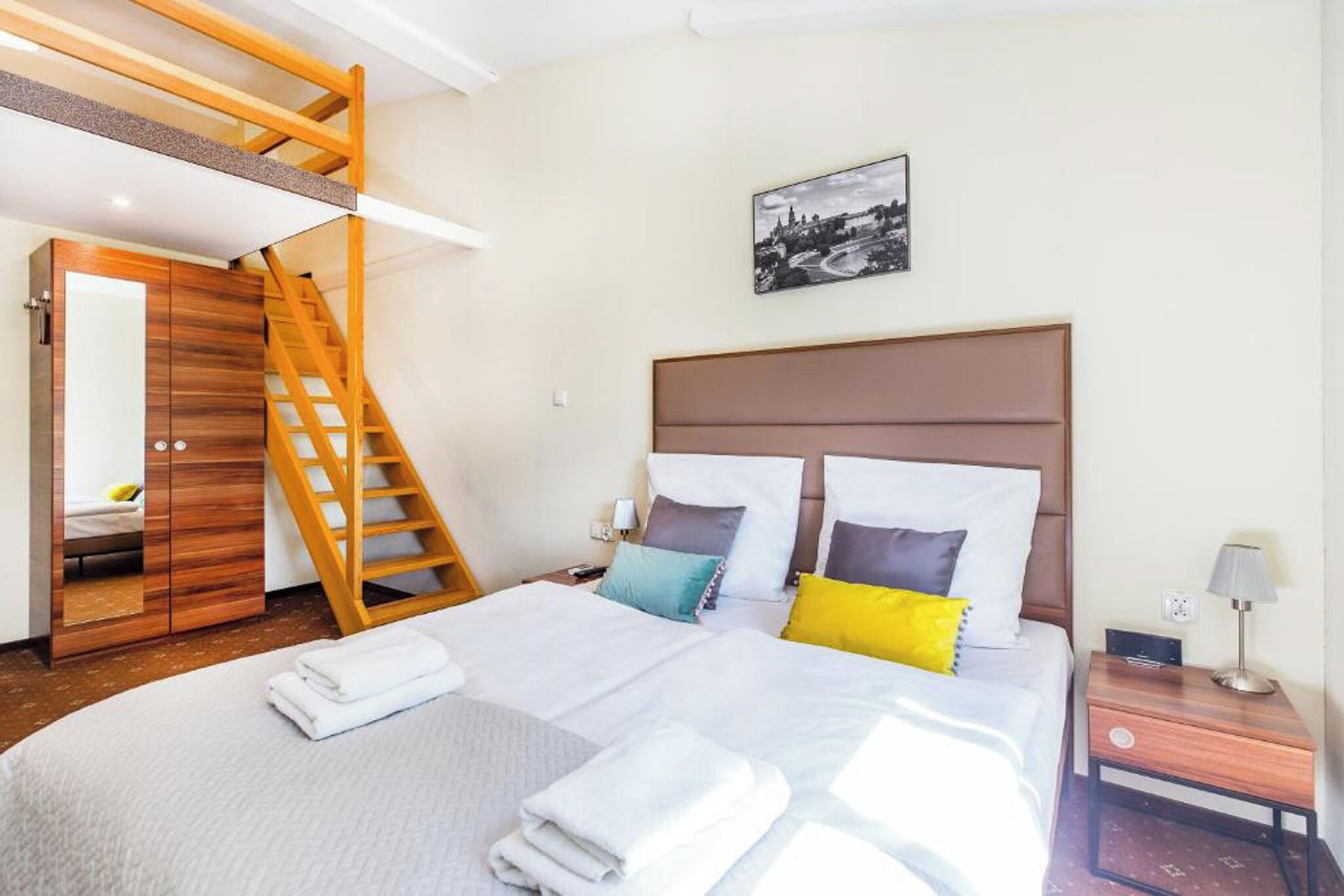 Quarto de hotel com cama, escada de madeira, mesa de cabeceira e quadro fixado na parede.