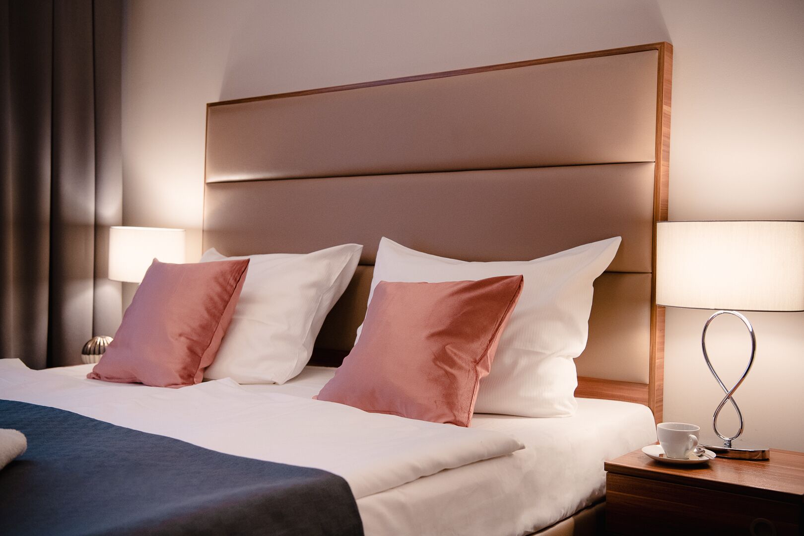 Una habitació d'hotel amb un llit gran, dues làmpades de nit, una taula de nit amb una tassa i un plat, i un capçal de llit.