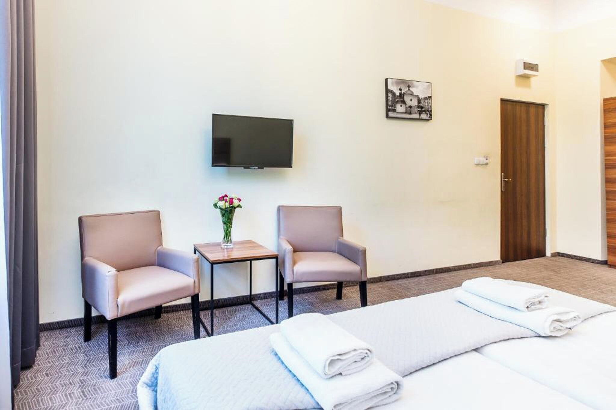 A hotel room with two single beds, a TV, two armchairs, a small table with a vase, and a door.