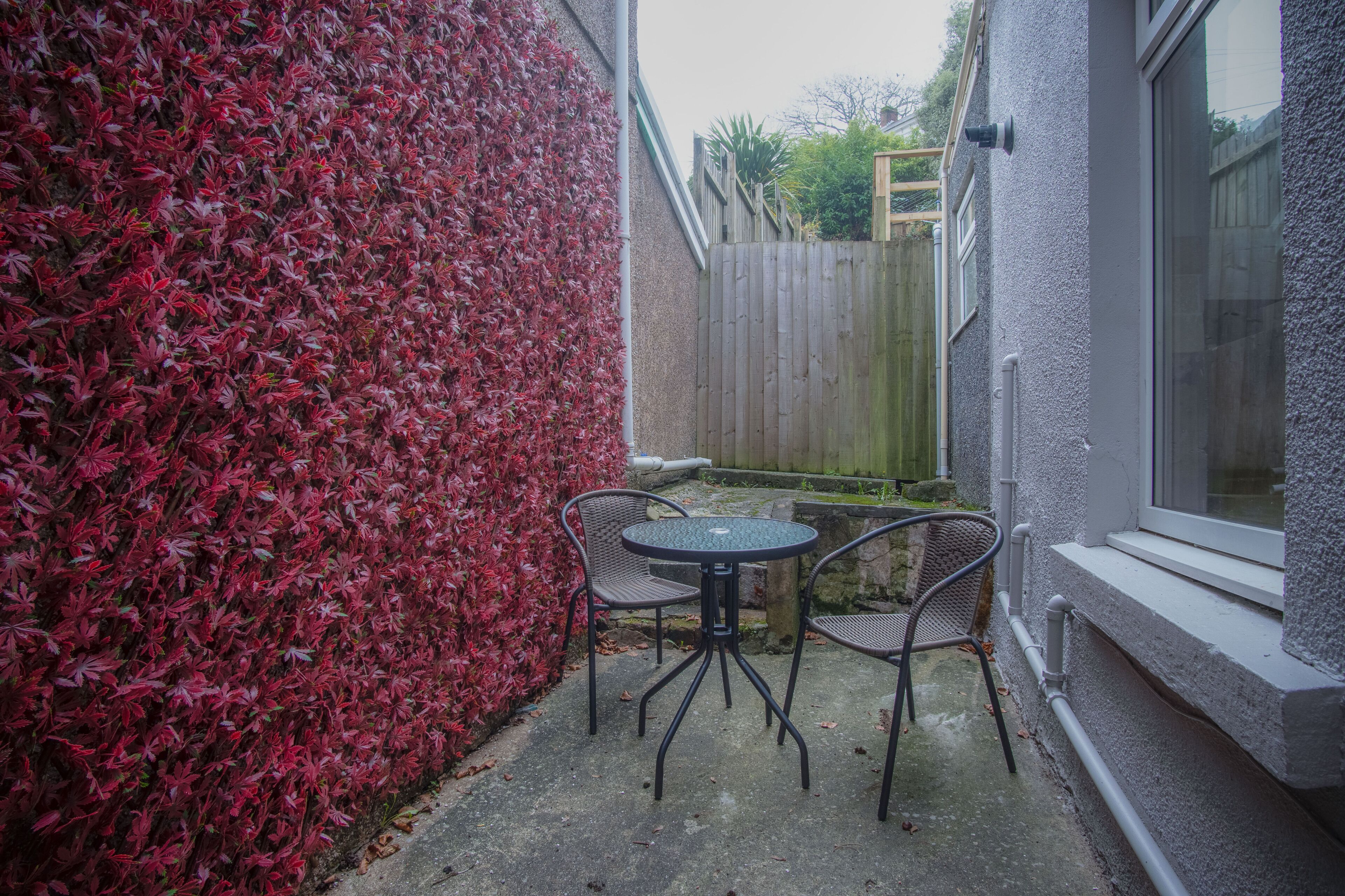 A patio with a round table and two chairs, a wooden fence, and a wall covered in red foliage.