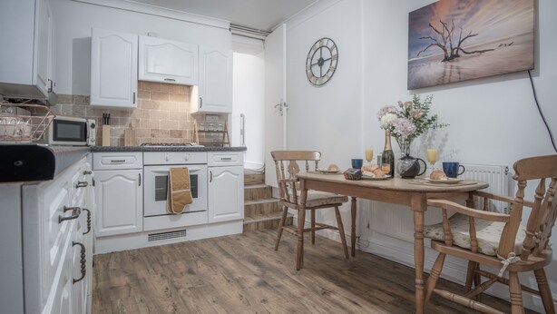 Una cocina con armarios blancos, una mesa de comedor de madera y sillas. Hay un reloj en la pared y un florero con flores sobre la mesa.