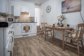 A kitchen with white cabinets, a wooden dining table, and chairs. There is a clock on the wall and a vase with flowers on the table. - Langland Road - 1 Bedroom - Mumbles (Swansea)