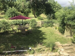 A patio area with a table and chairs under a red umbrella, surrounded by greenery and a stone wall. - Renovated wine cellar and outbuildings surrounded by nature (Villetritouls)