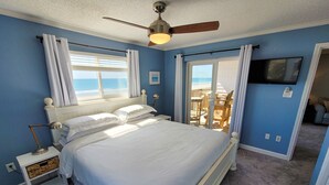A bedroom with a bed, a ceiling fan, a television, and a view of the beach.
