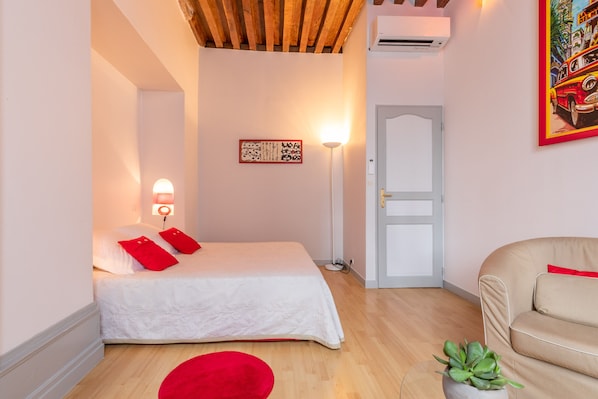 A bedroom with a bed, a red rug, a potted plant, a sofa, a wall-mounted lamp, and a door.