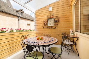 Een houten terras met een ronde tafel en stoelen, een decoratieve muur en een raam.
