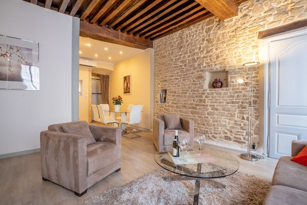 Un salon chaleureux avec un mur en pierre, deux canapés, une table basse en verre et, au fond, un coin repas.