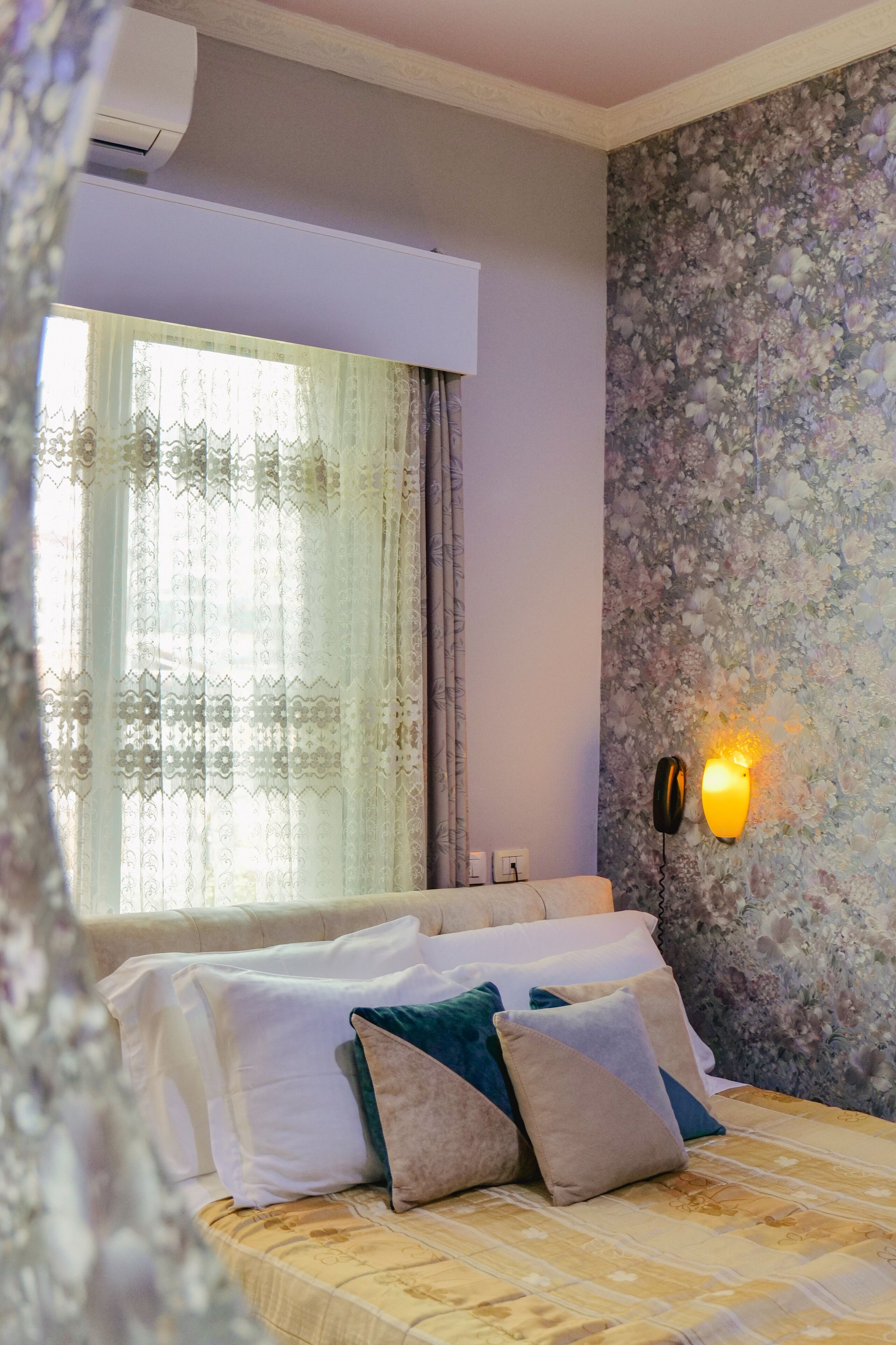 A bedroom with a bed, pillows, a window with curtains, a floral wallpaper, and a wall-mounted light fixture.
