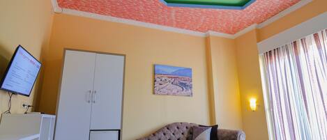 A hotel room with a green and orange ceiling, a bed with a striped bedspread, a sofa with a pillow, a flat-screen TV, and a painting on the wall.