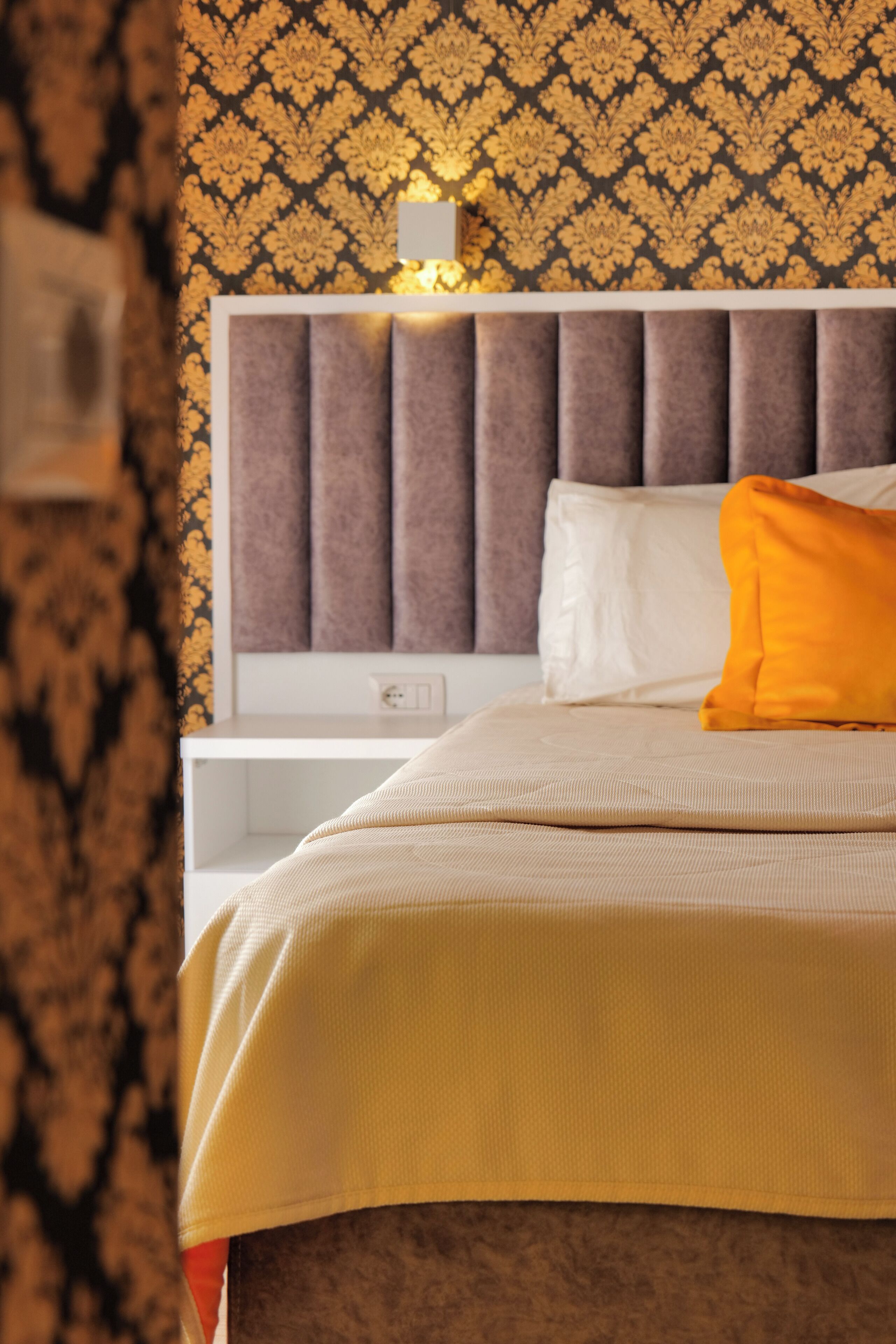 A neatly made bed with a white headboard, a beige bedspread, and a single orange pillow.