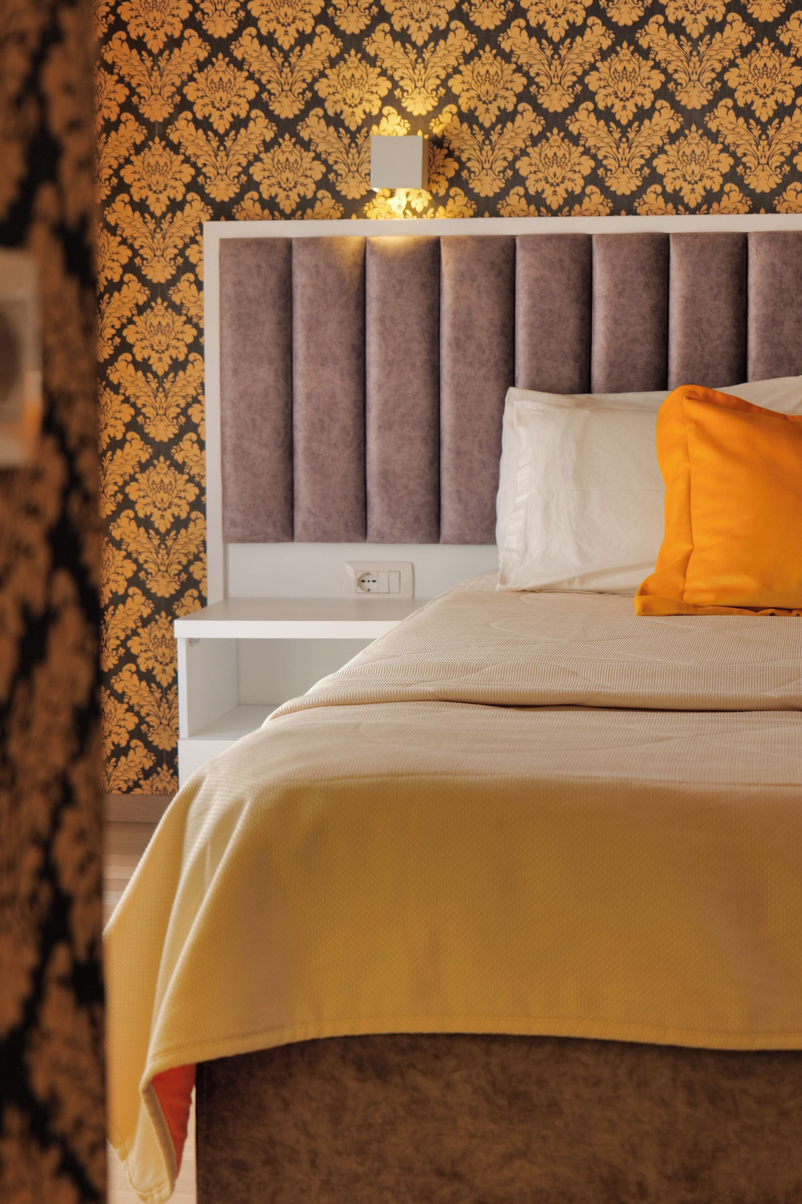 A neatly made bed with a headboard, a bedside table, and a wall with a patterned wallpaper.