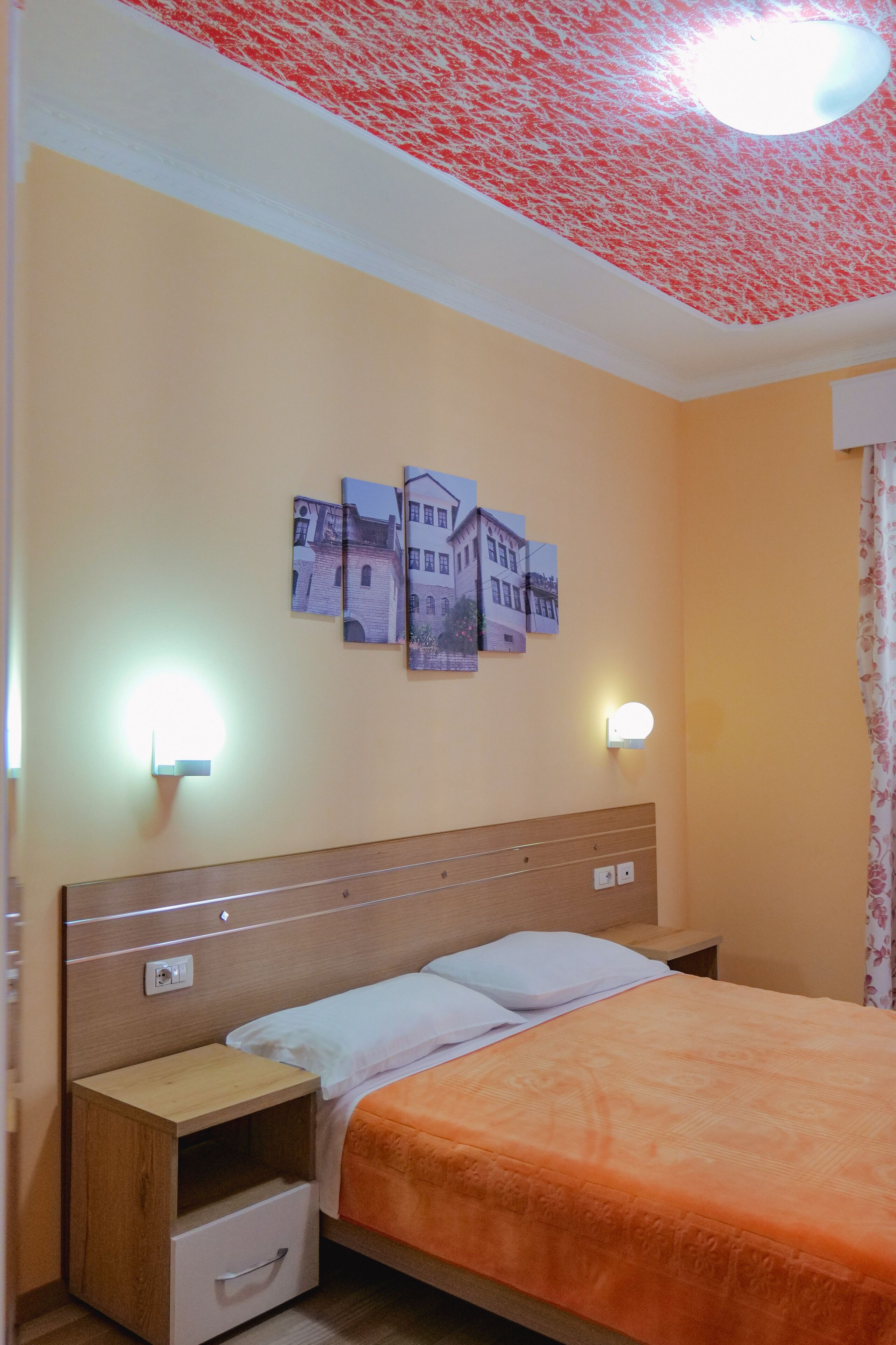 Un dormitorio con cabecera de madera, una cama individual con colcha naranja, una mesita de noche con cajón, y lámparas fijadas a la pared.