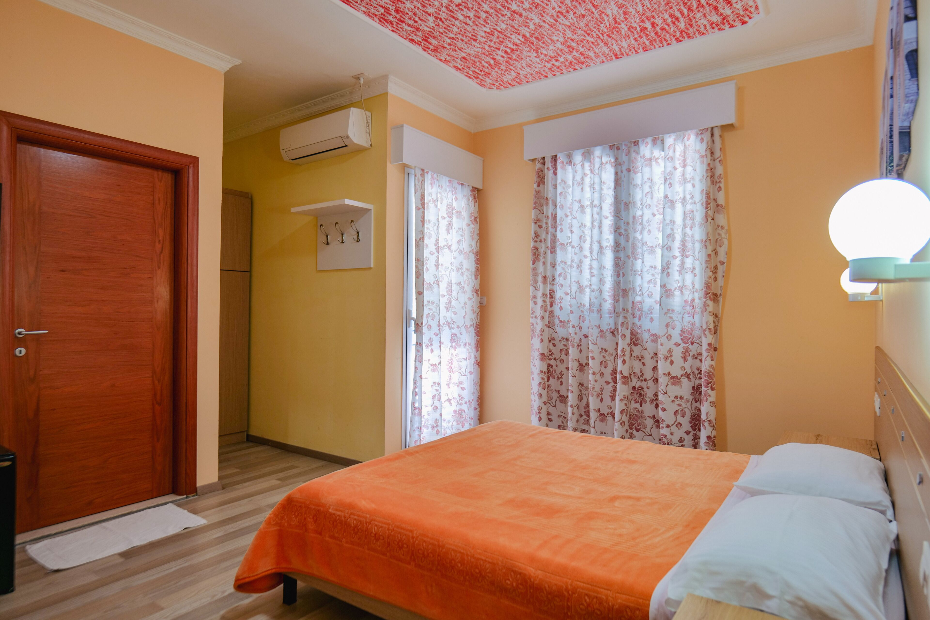 A bedroom with a wooden door, an air conditioning unit, a bed with an orange bedspread, and a wall-mounted light fixture.