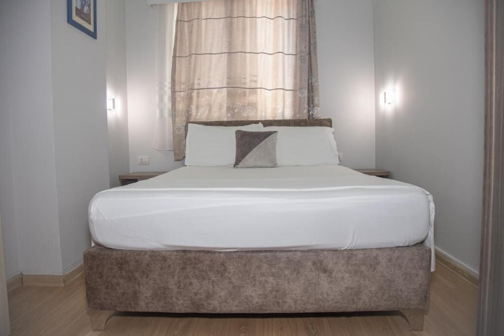 A bedroom with a bed, a nightstand, a window with curtains, and a wall-mounted light.