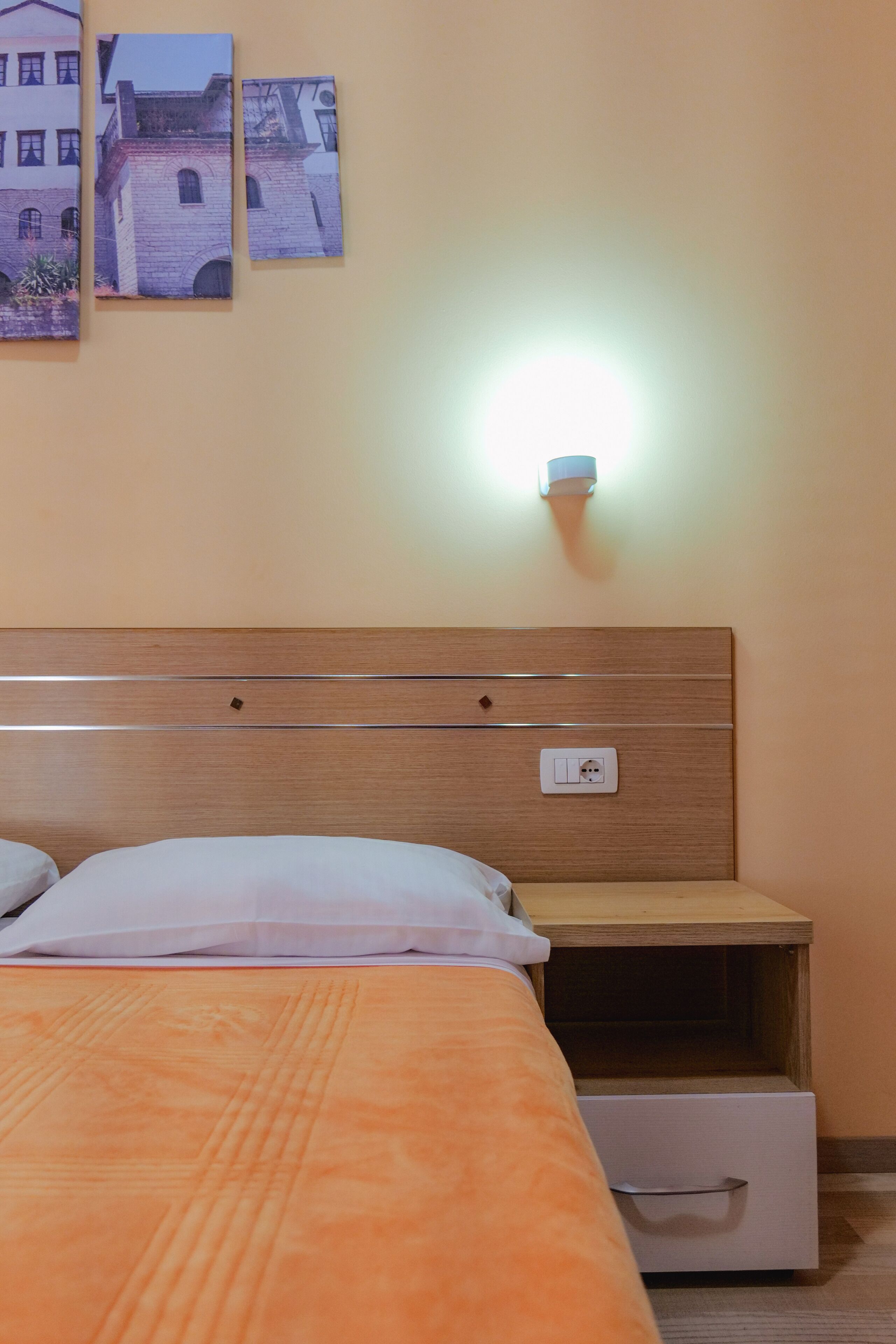 Habitación de hotel con cabecera de madera, cama con colcha naranja, mesita de noche con lámpara y aplique de pared.