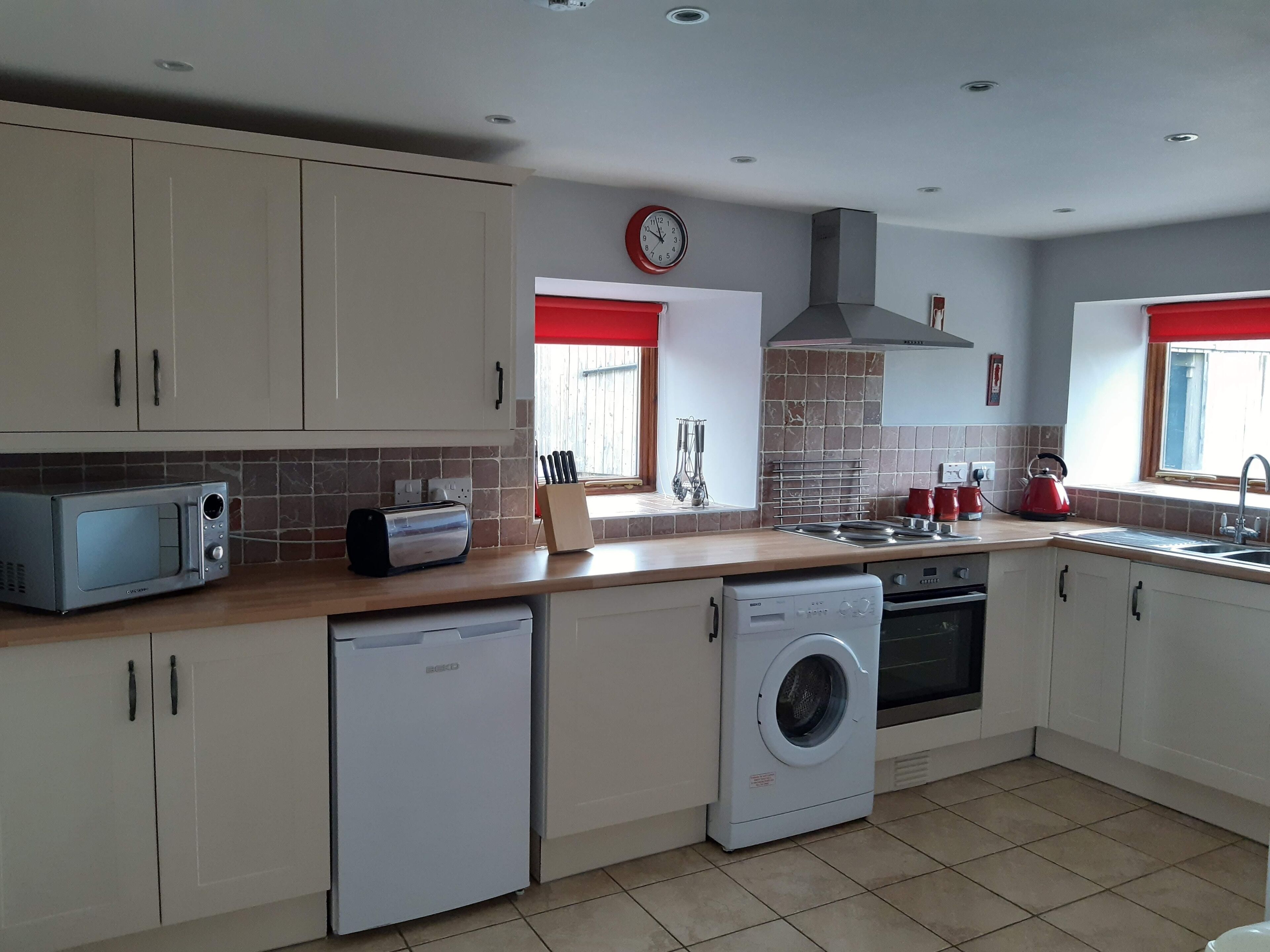 A modern kitchen with white cabinets, a microwave, a toaster, a dishwasher, a washing machine, a stove, and a red kettle.