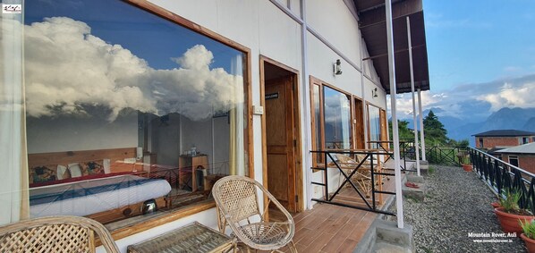 A room with a bed, a window, and a balcony with wicker chairs and potted plants. - Mountain Rover Auli (Joshimath)