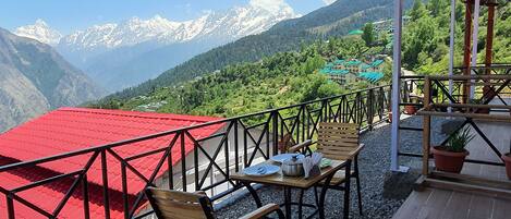 A terrace with a red roof, wooden chairs, a table with dishes, and a view of snow-capped mountains and a village.