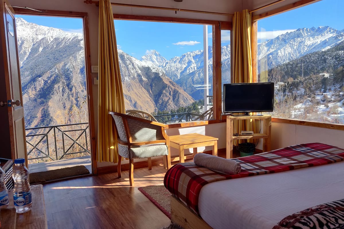 Ein Hotelzimmer mit Bett, Fernseher, Sessel, Tisch und Blick auf schneebedeckte Berge.