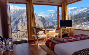 A hotel room with a bed, a TV, a chair, a table, and a view of snow-covered mountains. - Mountain Rover Auli (Joshimath)