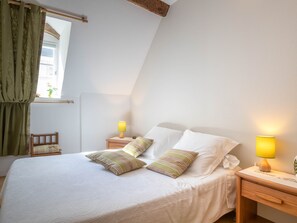 Una cama bien hecha con dos almohadas, una mesita de noche con lámpara y una ventana con cortinas.