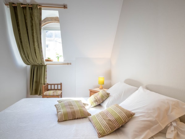 A neatly made bed with striped pillows, a bedside lamp, and a view of a building through a window with green curtains.