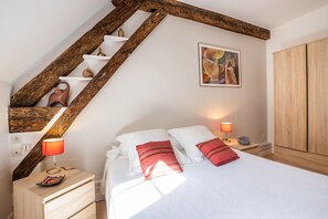 Un dormitorio con una escalera de madera, una cama con ropa de cama blanca, dos almohadas rojas, una mesita de noche con una lámpara y un cuadro en la pared.