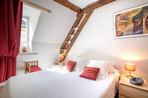 A bedroom with a sloped ceiling, a wooden ladder, a bed with white and red bedding, a bedside table with a lamp, and a painting on the wall.