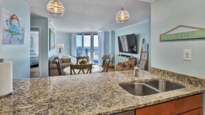 Una cocina moderna con encimeras de granito y electrodomésticos de acero inoxidable, un comedor con una mesa redonda y sillas, y una sala de estar con televisor y vista al océano.