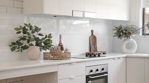 Una cucina moderna con mobili bianchi, forno integrato e un piano di lavoro con oggetti decorativi, tra cui una pianta, una bottiglia e un tagliere di legno.