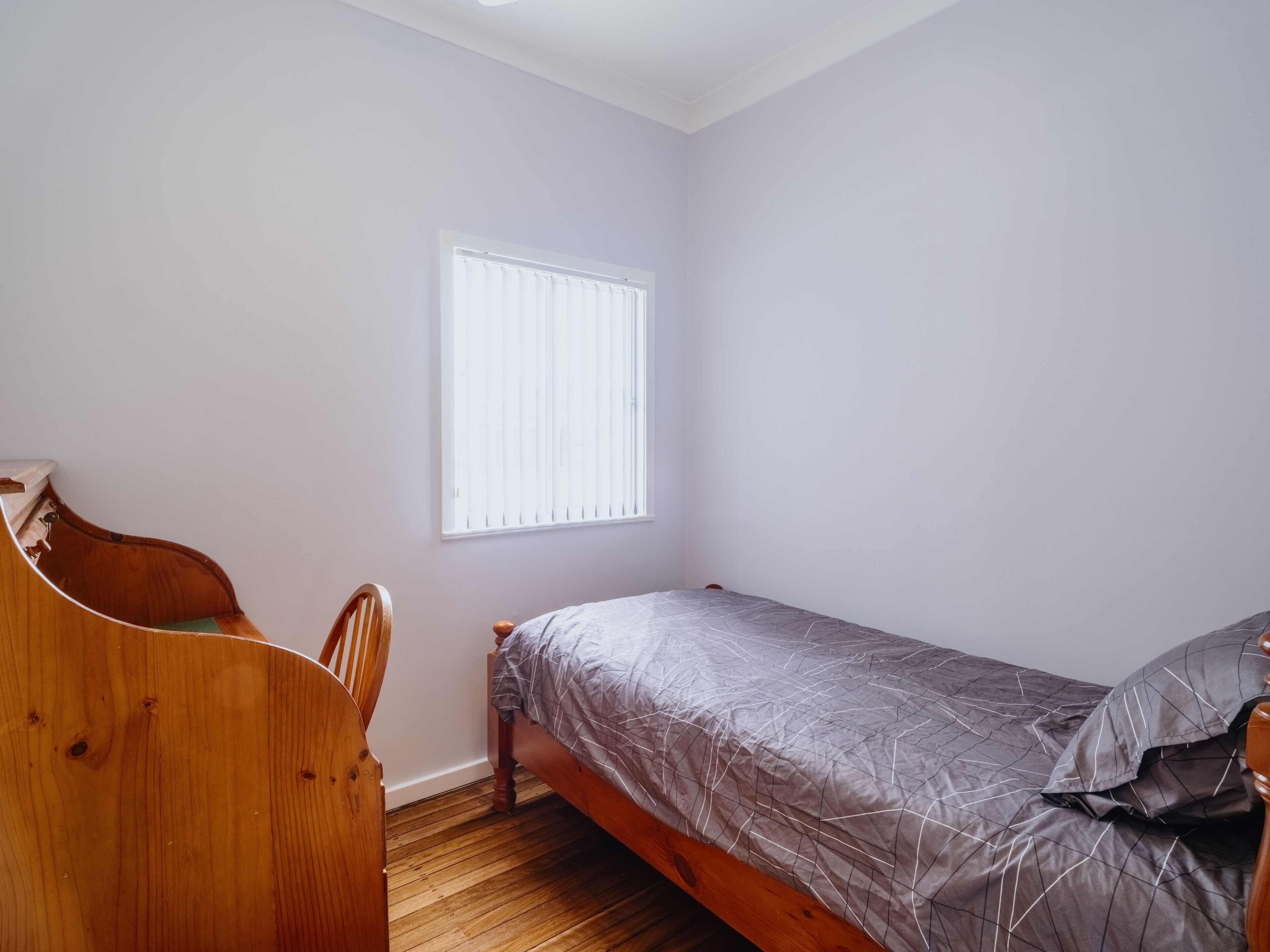 Ein kleines Zimmer mit einem Einzelbett, einem Holzstuhl, einem Fenster mit Jalousien und Holzboden.