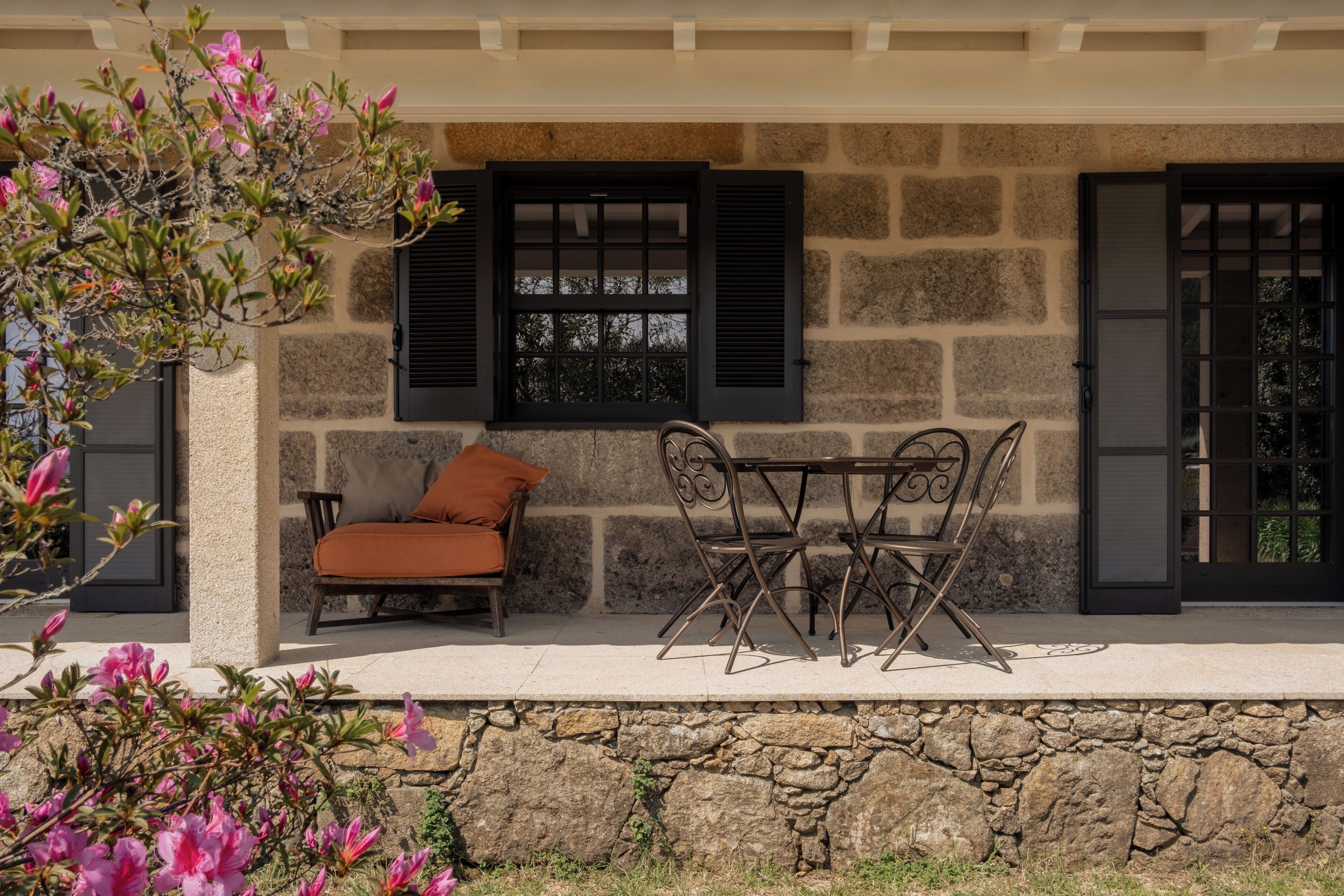 Pared exterior de piedra con una ventana y una puerta, un juego de mesa y sillas, y plantas con flores.
