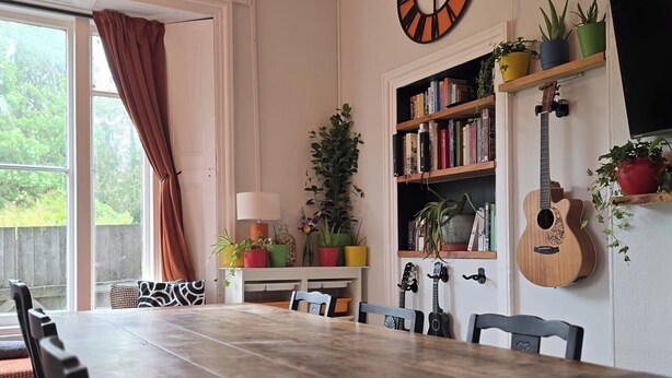 A dining area with a wooden table, chairs, a clock, bookshelves, a guitar, and potted plants. - Spacious 2 Bed/Bath Central Apt, Parking & Garden (Exeter)