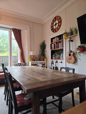 Un coin repas avec une table en bois, des chaises, une horloge, des étagères à livres, une guitare et des plantes en pot.
