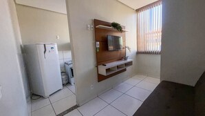 A compact kitchen with a refrigerator, a gas cylinder, a TV mounted on a wooden shelf, and a window with blinds. - CONDOMÍNIO SONHARE - 102 (Anapolis)