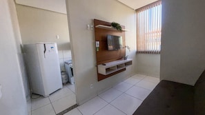 A compact kitchen with a refrigerator, a gas cylinder, a TV mounted on a wooden shelf, and a window with blinds. - CONDOMÍNIO SONHARE - 102 (Anapolis)