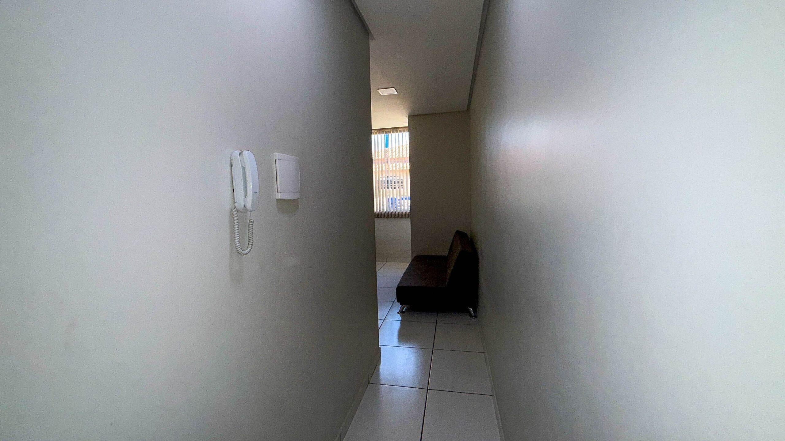 Un couloir étroit avec un téléphone fixé au mur, une chaise et une fenêtre avec des stores.