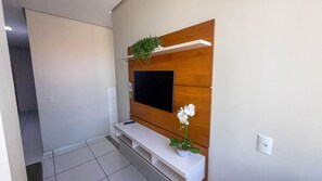 A modern living room with a flat-screen TV mounted on a wooden panel, a small white console table, and a potted orchid. - CONDOMÍNIO SONHARE - 102 (Anapolis)