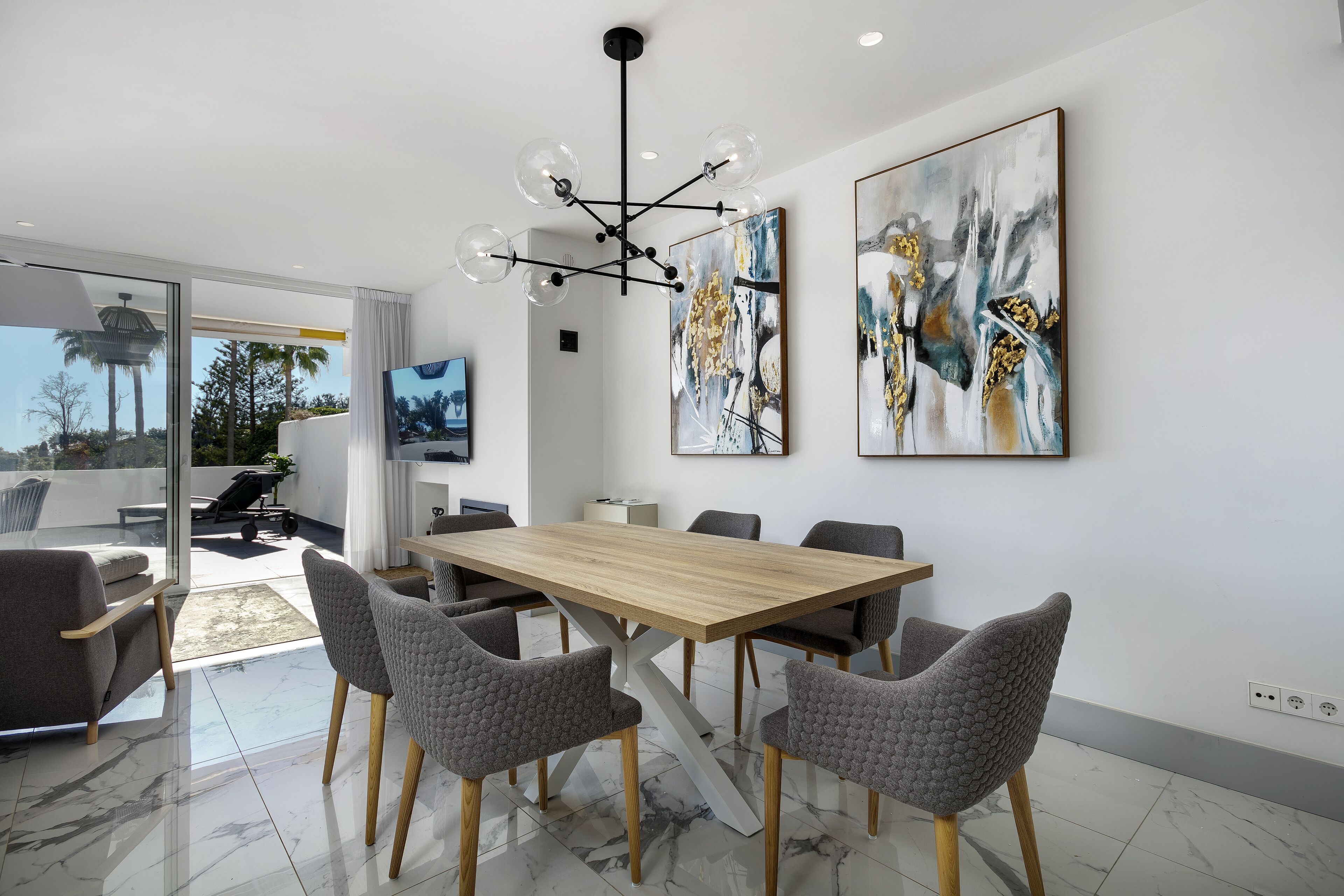 A modern dining area with a wooden table and grey upholstered chairs, a large abstract painting on the wall, and a view of the outdoors through a glass door.
