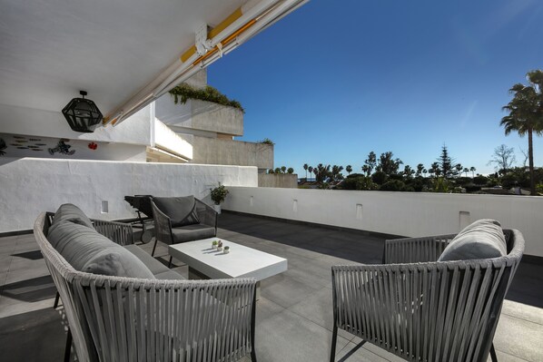 Una terraza en la azotea con un área de estar moderna al aire libre, que incluye un sofá, sillas, una mesita y una planta en maceta.