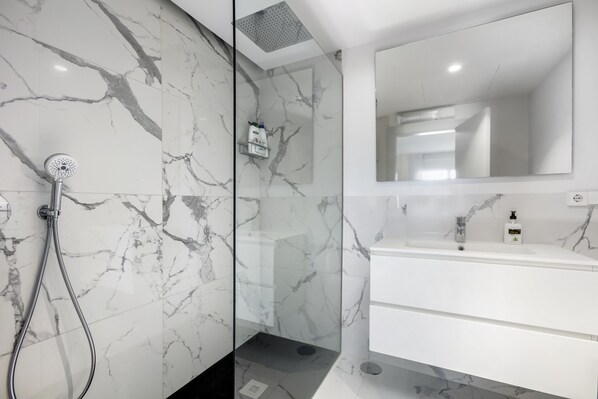 Une salle de bain moderne avec une douche en verre, des murs en marbre, un grand miroir et un meuble-lavabo blanc.
