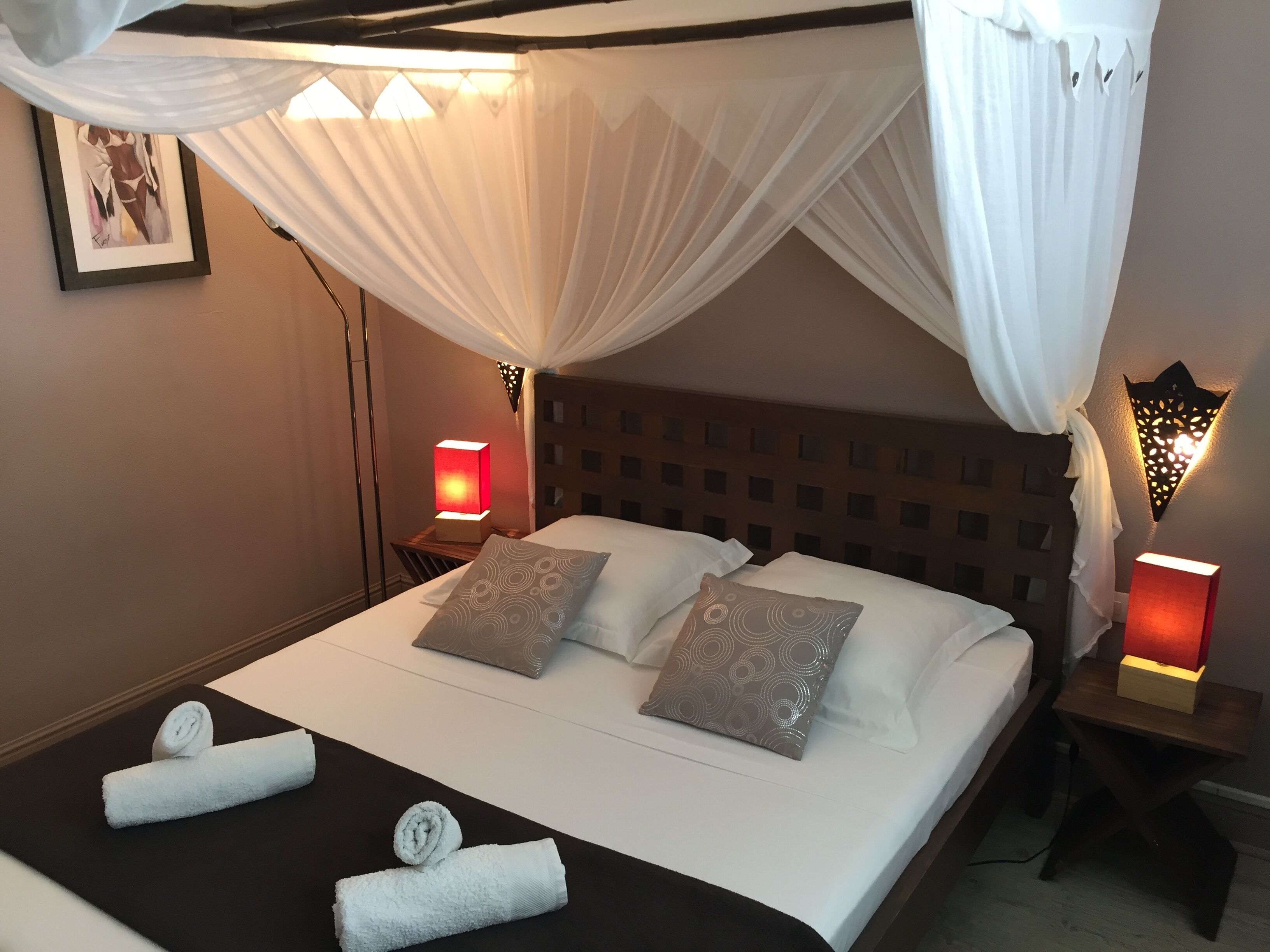 Een net opgemaakt bed met witte lakens, twee kussens en twee opgerolde handdoeken. Twee bedlampen met rode kapjes, een wandlamp en een lijst met een afbeelding aan de muur.