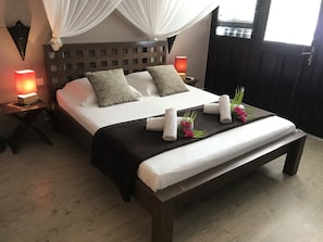 A neatly made bed with a headboard, two bedside tables with lamps, a bench at the foot of the bed, and a window with curtains.