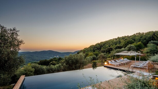 Une piscine à débordement avec une terrasse en bois, des chaises longues et des parasols, le tout entouré d’une végétation luxuriante et de collines.