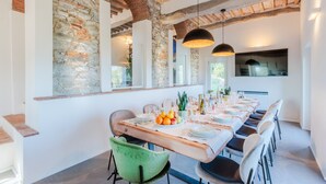Un espace repas avec une longue table dressée pour accueillir plusieurs personnes, des murs en pierre, des poutres en bois et des luminaires suspendus.